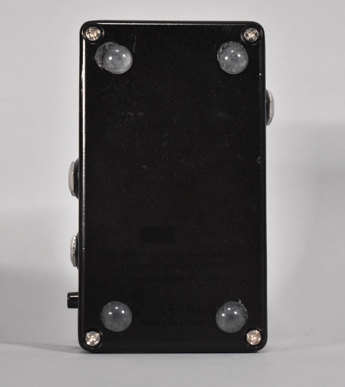 The black pedal is viewed from the bottom, showing four clear rubber feet and four silver screws, with minor scuffs.