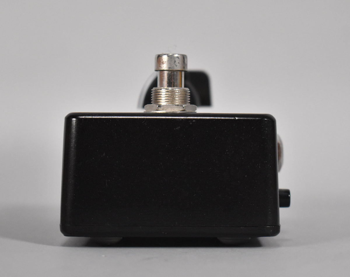Side view of the black boost pedal, showing a silver footswitch on top and a small black jack on the right side.