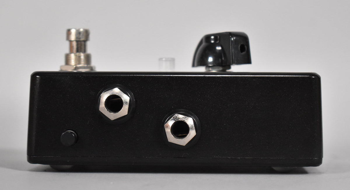 The black pedal is viewed from the side, showing a footswitch, a black knob, two input jacks, and a small black button.