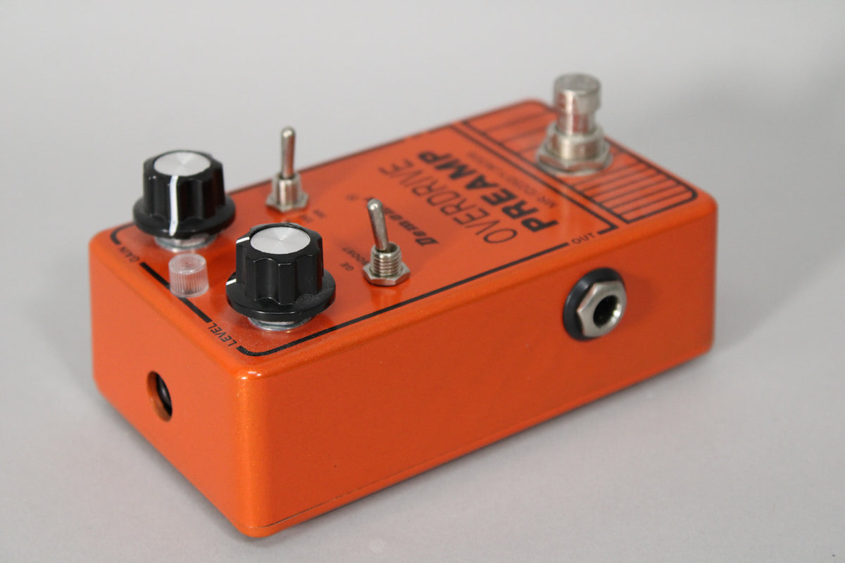 An orange metallic pedal, angled from the front right, showing two black knobs, three silver toggles, and an output jack.