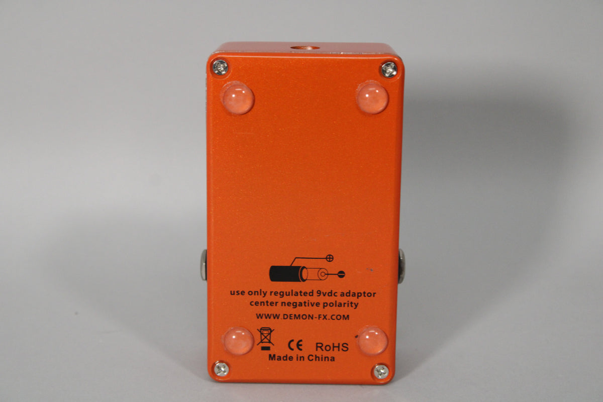 The orange pedal is viewed from the bottom, showing four clear rubber feet, four silver screws, and power requirements.