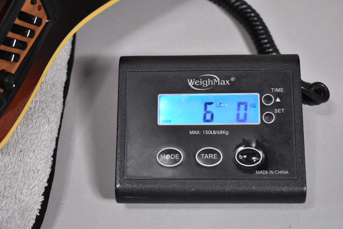 The guitar's sunburst body rests on a white towel, with a digital scale displaying "6 LB 0 OZ" in the foreground.