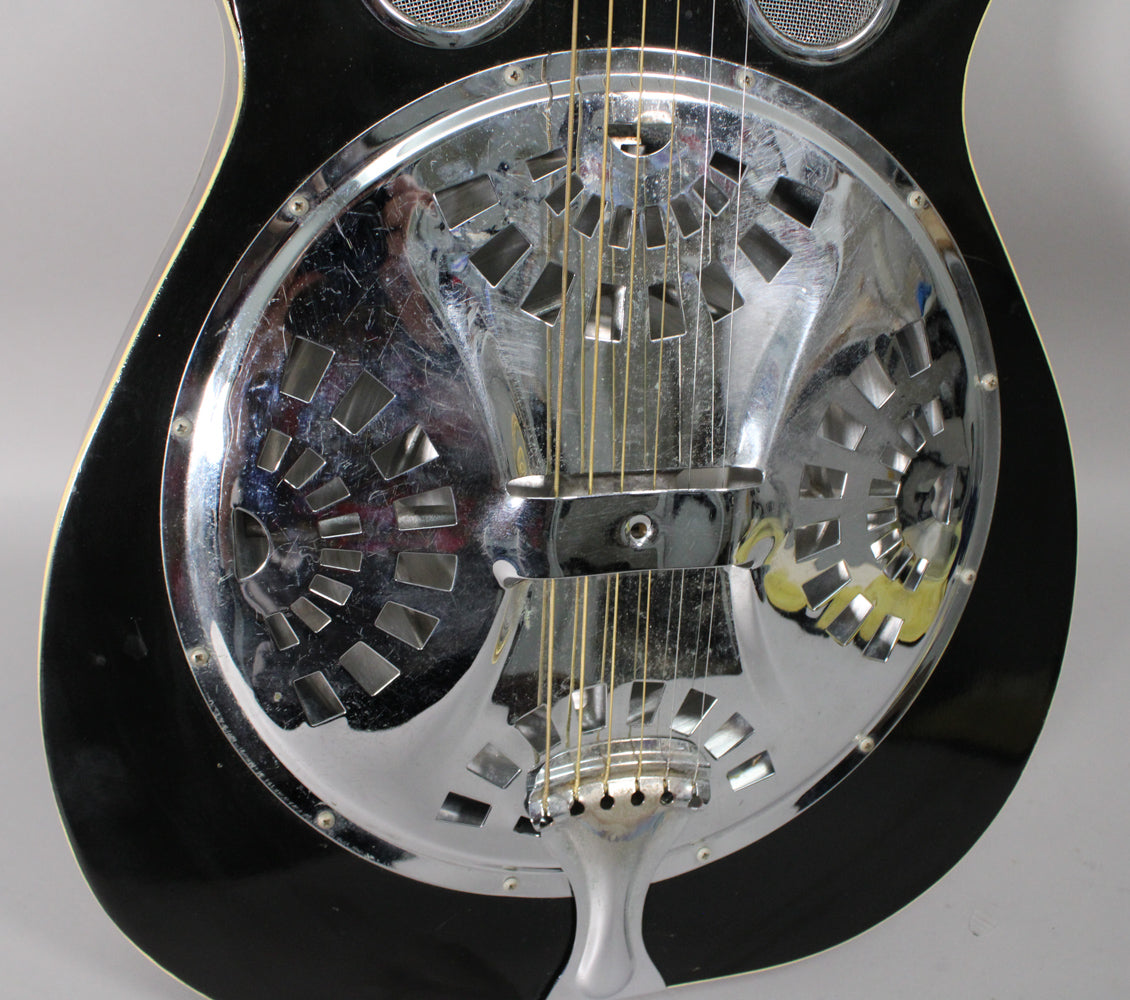 Close-up of the black resonator guitar's chrome cone with geometric cutouts, showing light scratches and a chrome tailpiece.