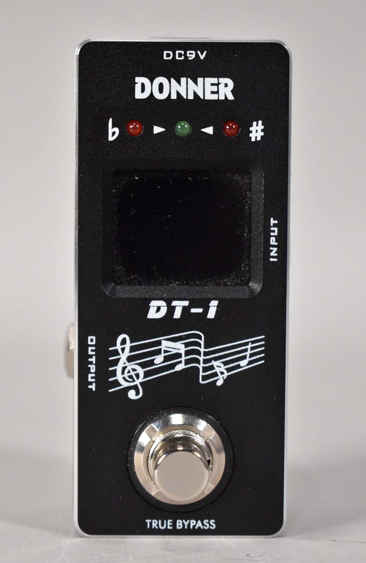 A black mini tuner pedal, viewed from the front, with a silver footswitch, a small screen, and red and green indicator lights.