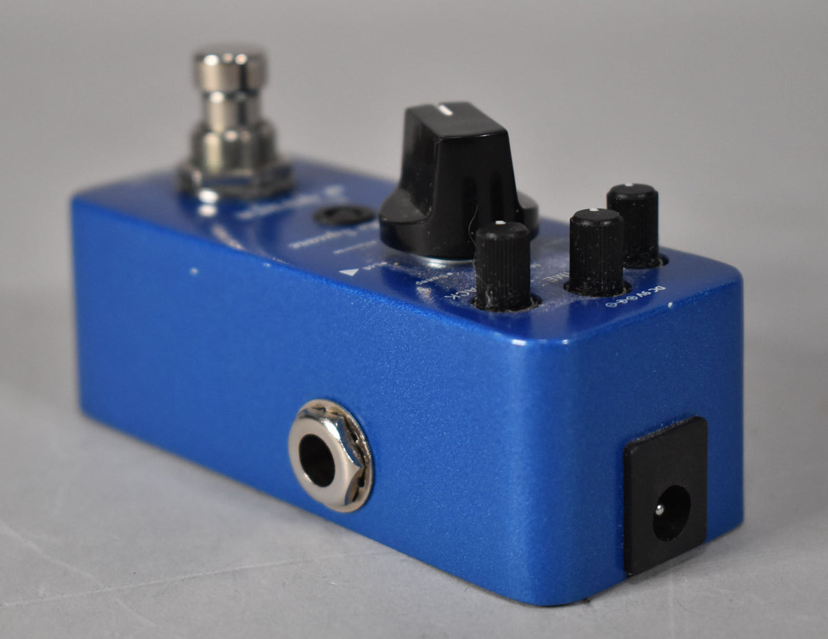 A blue delay pedal, angled from the left, showing a large black knob, three smaller black knobs, and input/power jacks.