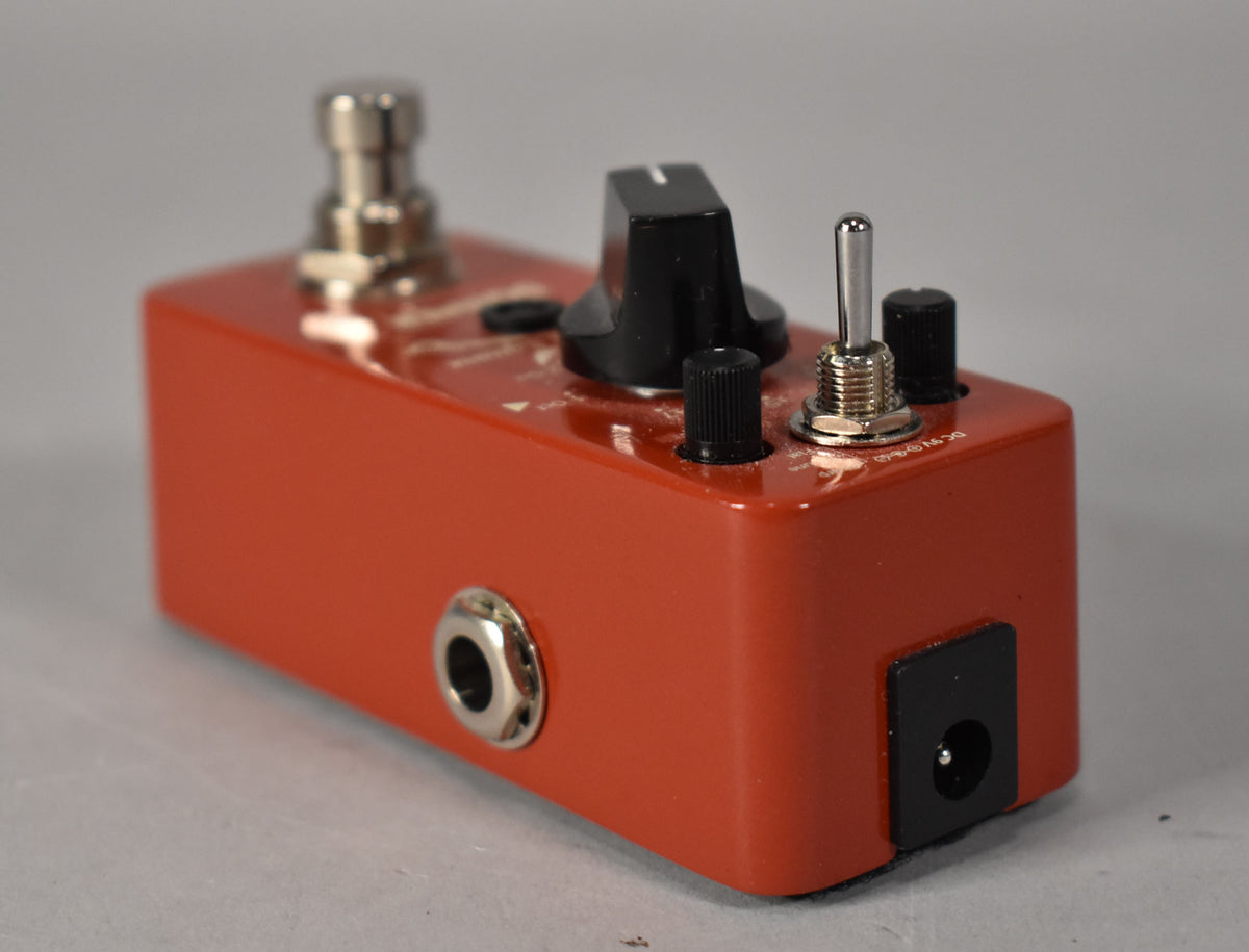 A reddish-orange pedal, viewed from the right, showing a chrome input jack, a large black knob, and a toggle switch.