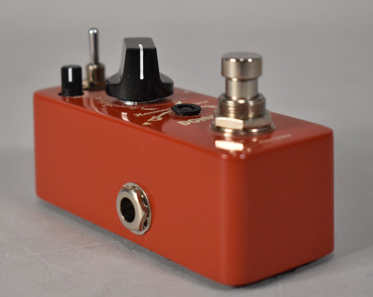 A bright orange pedal, viewed from the front-right, showing a large black knob, small black knob, toggle switch, and footswitch.