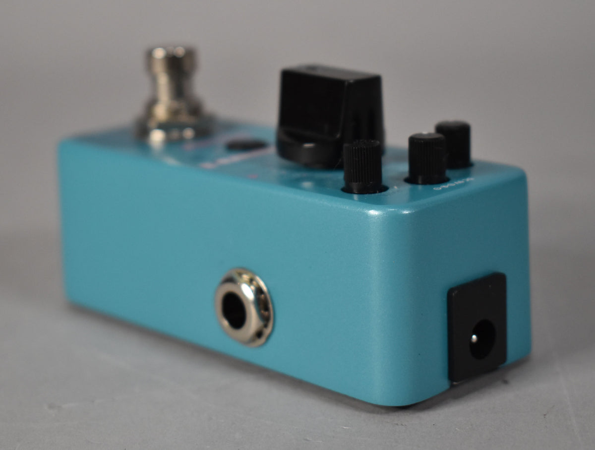 A turquoise pedal, viewed from the right side, showing a chrome jack, black power input, and three black knobs on top.