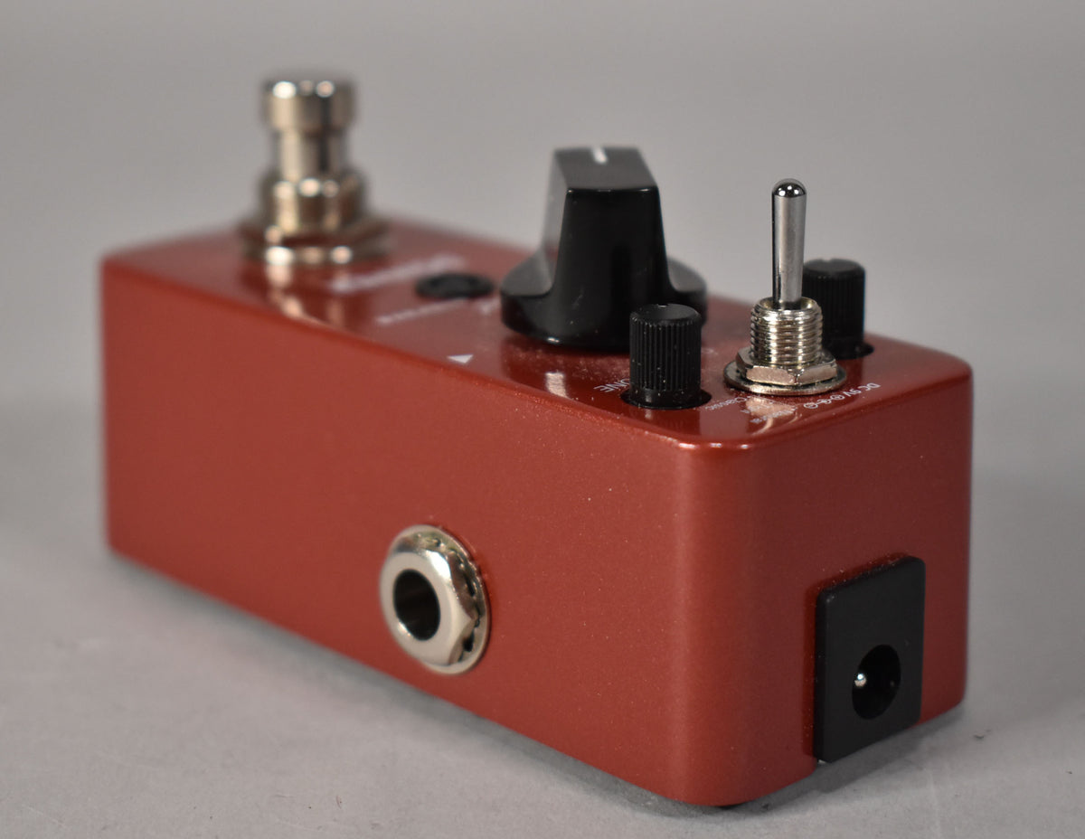 A reddish-brown pedal, angled from the right, showing a large black knob, two small black knobs, a toggle switch, and input jacks.