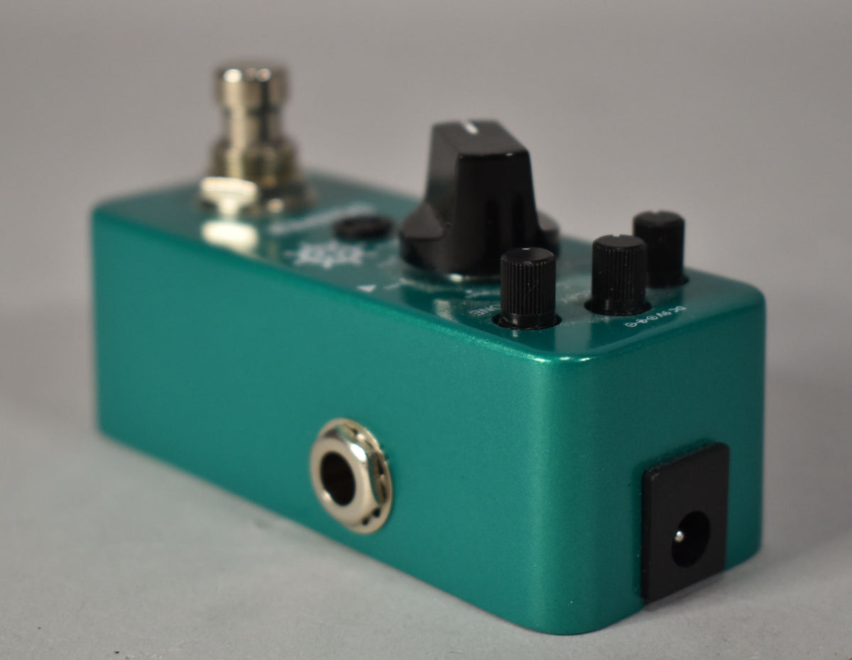 The teal metallic pedal is angled from the left, showing a large black knob, three smaller black knobs, and input/output jacks.