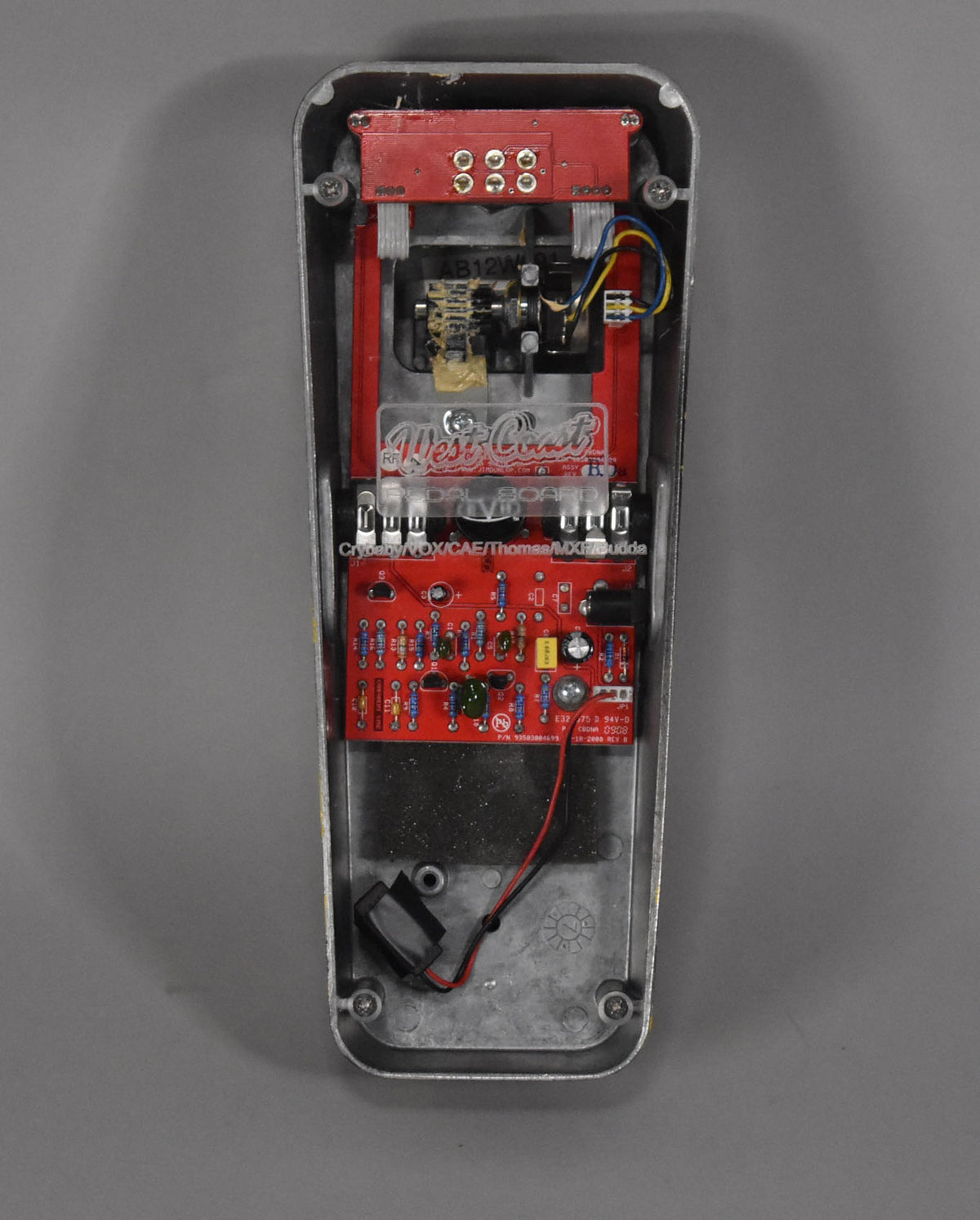 The wah pedal's bottom cover is removed, revealing red circuit boards, wiring, and internal components within the silver casing.
