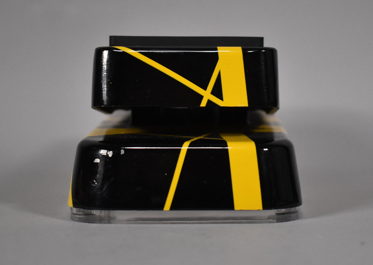 The wah pedal is viewed from the front, showing its glossy black finish with yellow stripes and some minor paint chips.