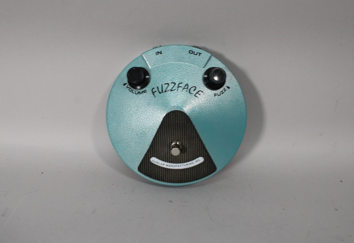 A round, teal-colored fuzz pedal with a textured finish, two black control knobs, a silver footswitch, and a black textured footpad.