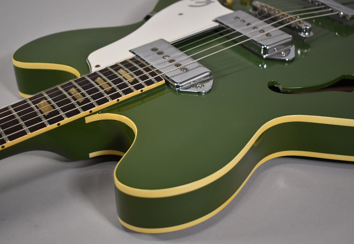 Close-up of the olive drab hollowbody guitar's lower bout, showing the neck, white pickguard, chrome pickups, and f-hole.