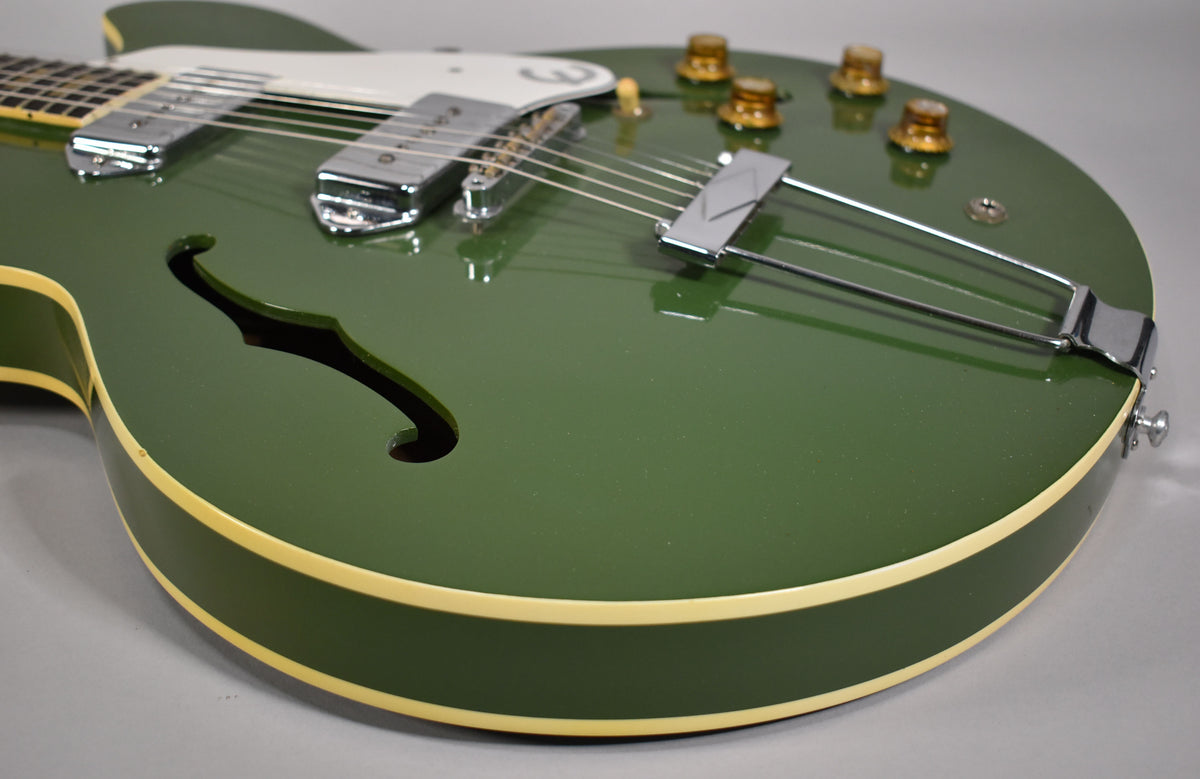 Upper body of a dark olive green hollowbody guitar, showing two chrome pickups, f-hole, and four amber control knobs.