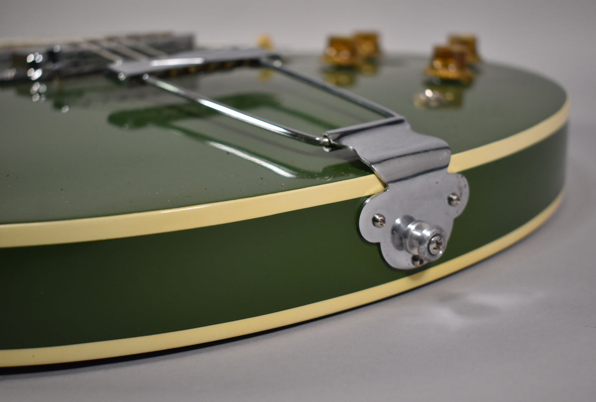 Close-up of the olive drab guitar body's lower bout, showing the chrome trapeze tailpiece, cream binding, and output jack.