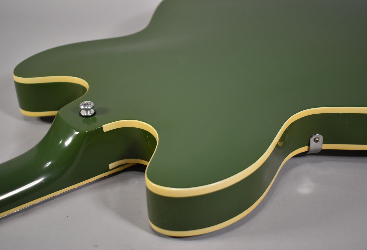 The guitar's olive drab body, with cream binding, is shown from the back, revealing a strap button near the neck heel and a small metal clip.