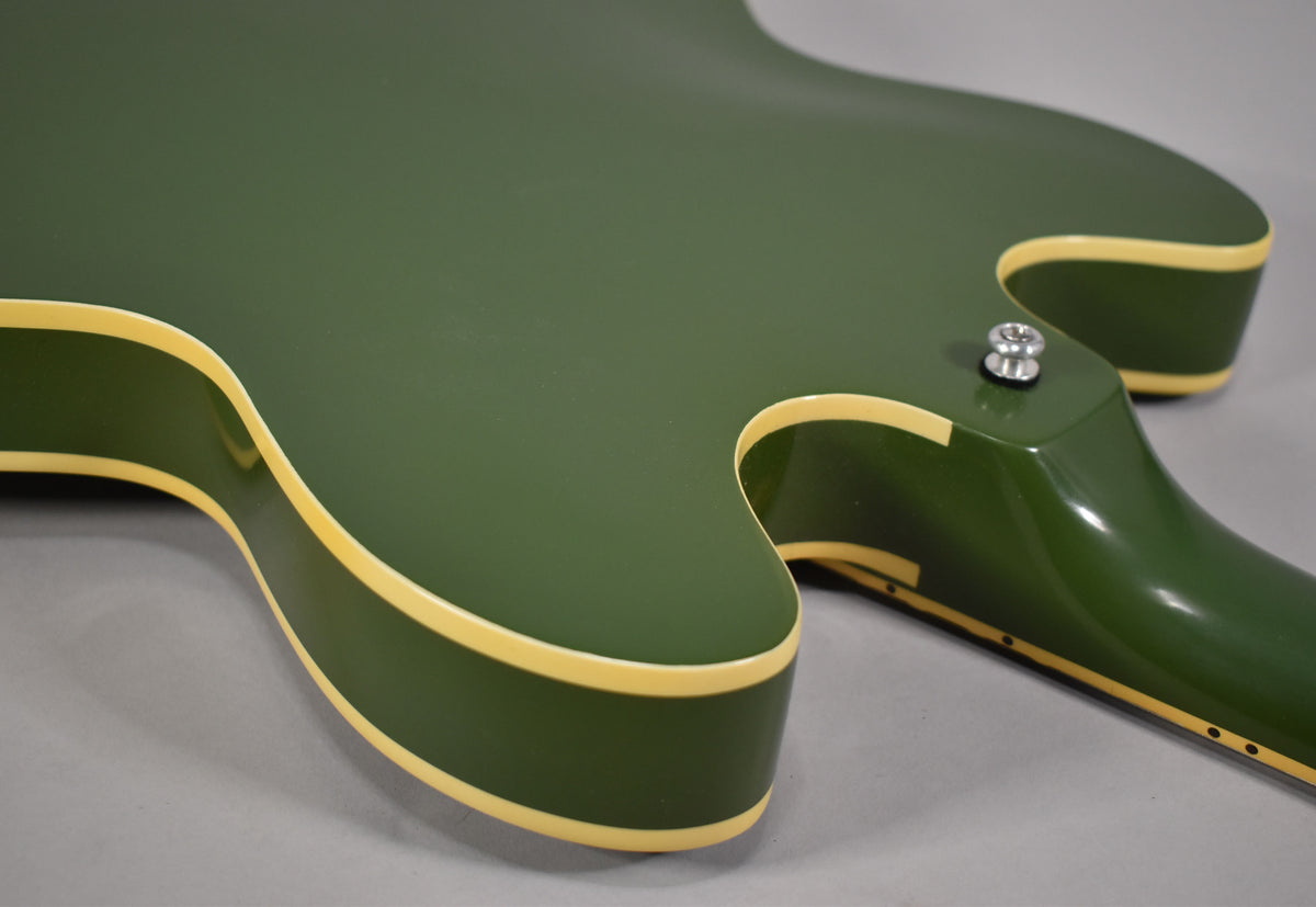 The guitar's olive drab body and neck are shown from the back, with cream binding visible along the edges and a strap button near the neck joint.