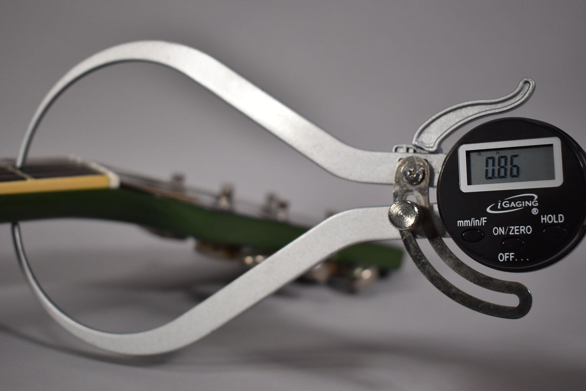 A digital caliper measures the olive drab headstock at 0.86 inches, with tuning pegs visible in the blurry background.