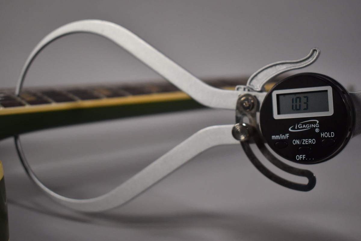 A digital caliper measures the guitar's fretboard, displaying "1.03" on its screen, with the olive drab finish visible.