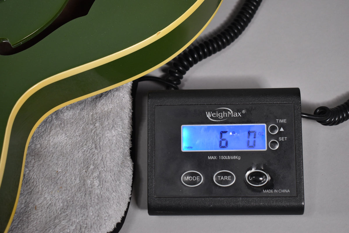 The olive drab hollowbody guitar's lower bout with cream binding rests on a grey bag, next to a scale displaying "6.0" lbs.