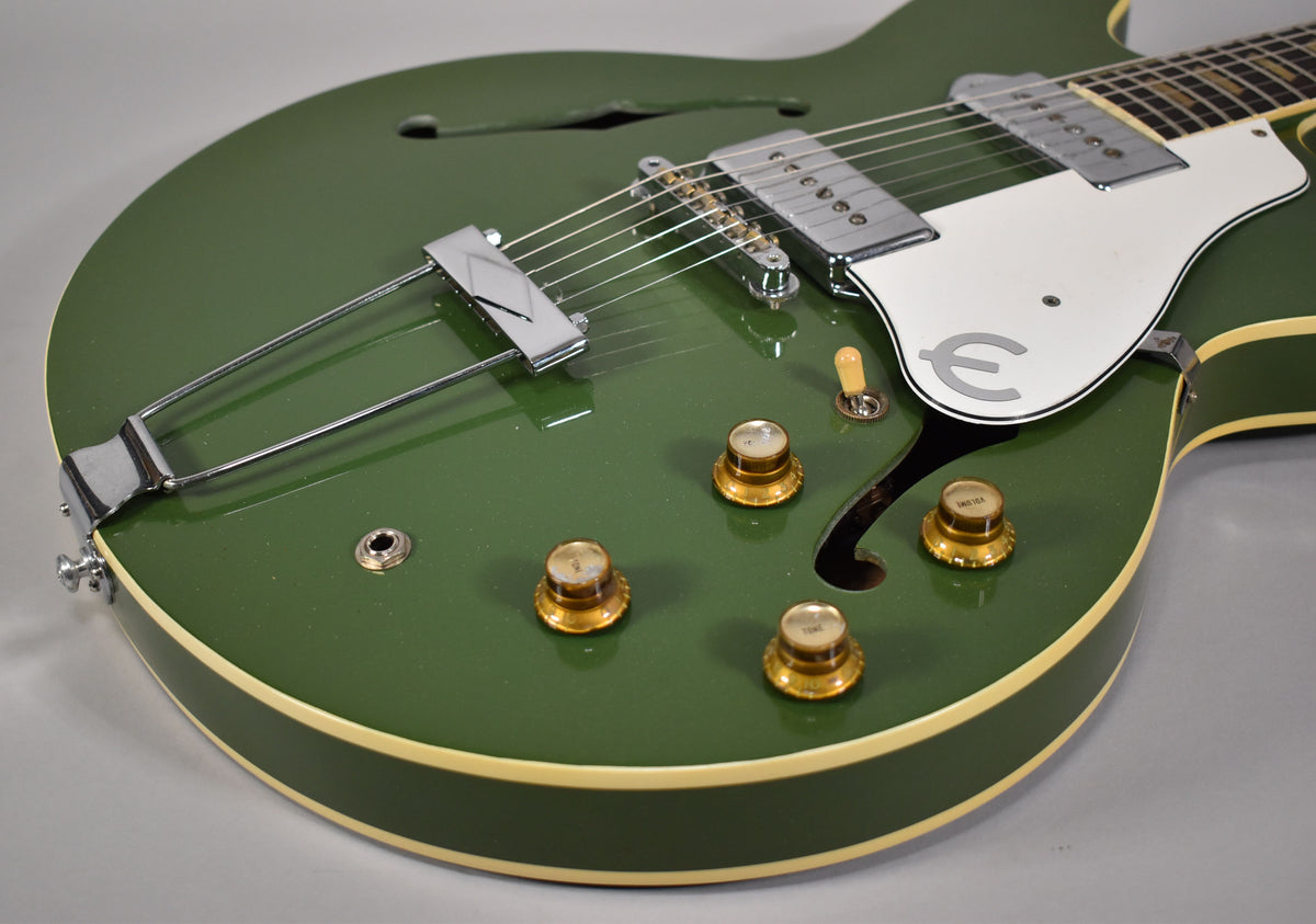 Upper body of a glossy olive drab hollowbody guitar, showing a trapeze tailpiece, two chrome pickups, and four gold knobs.