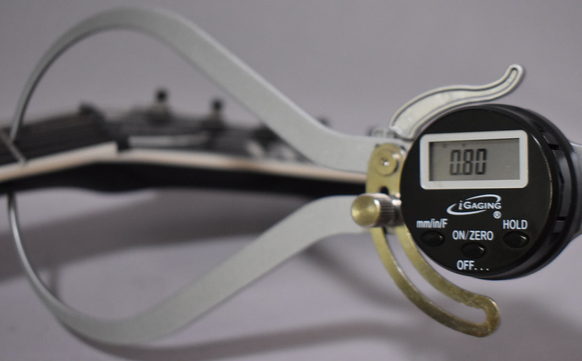 A digital caliper measures the guitar's nut, showing 0.00 on its screen, with the headstock blurred in the background.