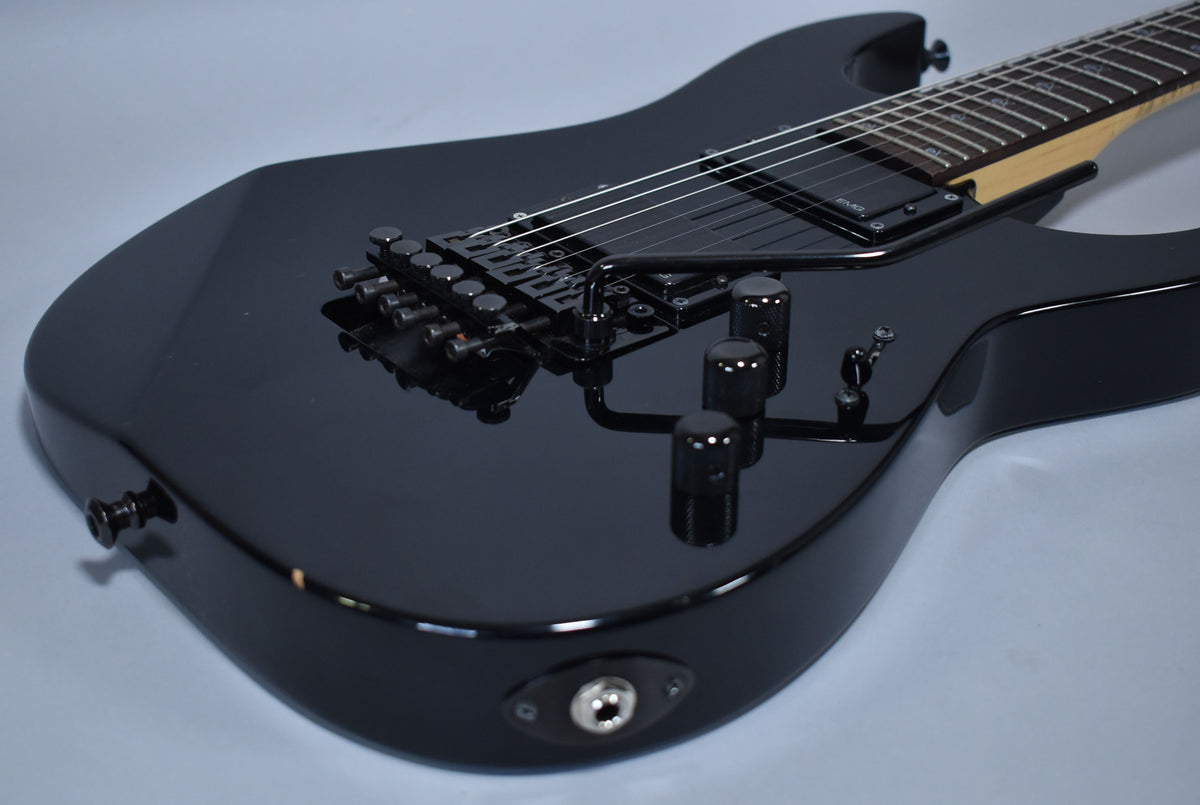The glossy black electric guitar body is angled from the lower right, showing the black Floyd Rose tremolo, three control knobs, and output jack.