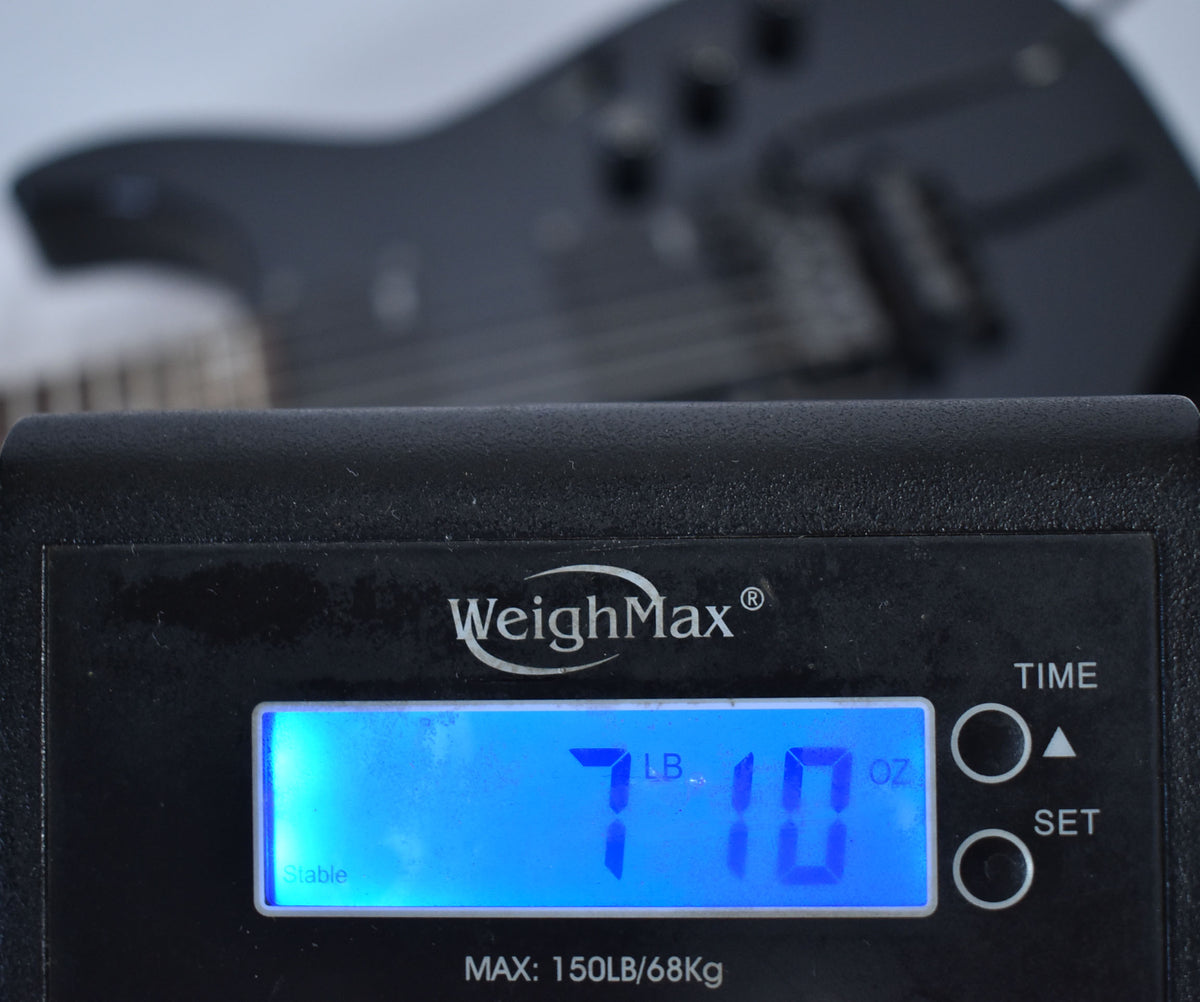 A black digital scale displays "7 LB. 10 OZ." with the guitar's black body and hardware blurred in the background.