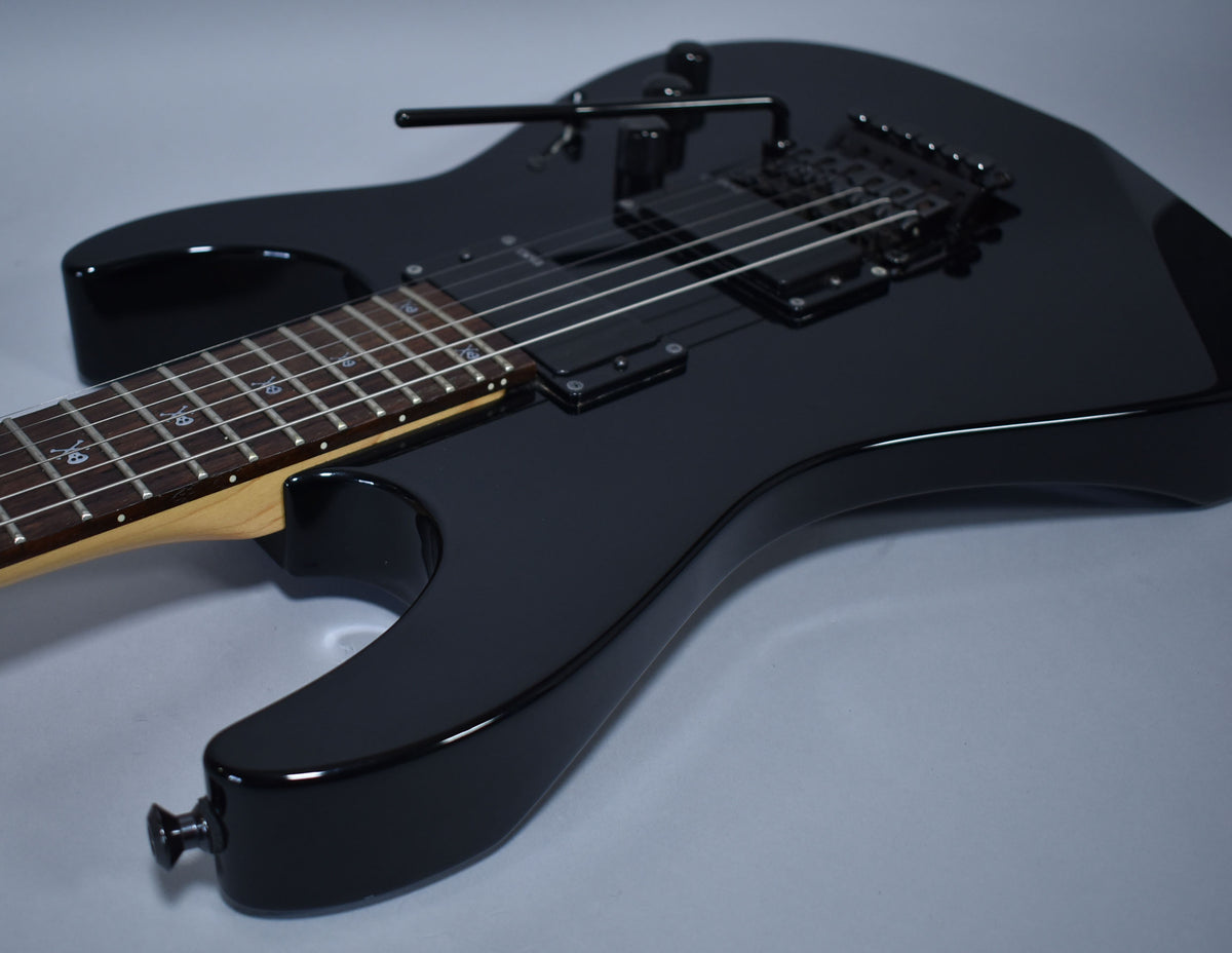 The black electric guitar, angled from the lower bout, shows its glossy finish, black hardware, and skull-and-crossbones fret inlays.
