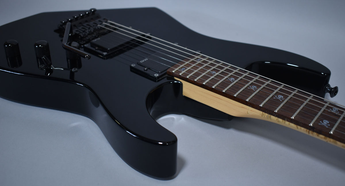 A glossy black electric guitar, angled from the lower bout, shows skull and crossbones fret markers and black hardware.