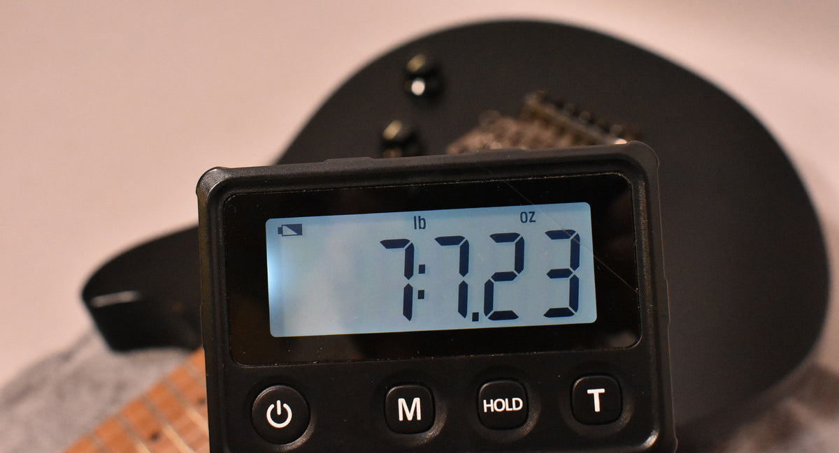 A digital scale displaying "7:7.23 lb oz" is centered, obscuring most of the satin black guitar body and its two black control knobs.