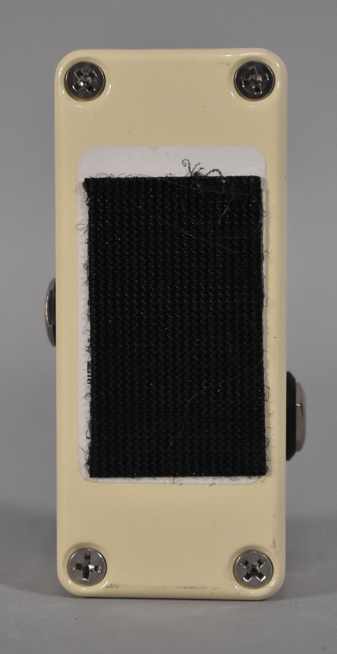 The bottom of the cream pedal is shown, with four silver Phillips head screws and a large black velcro strip attached to a white label.