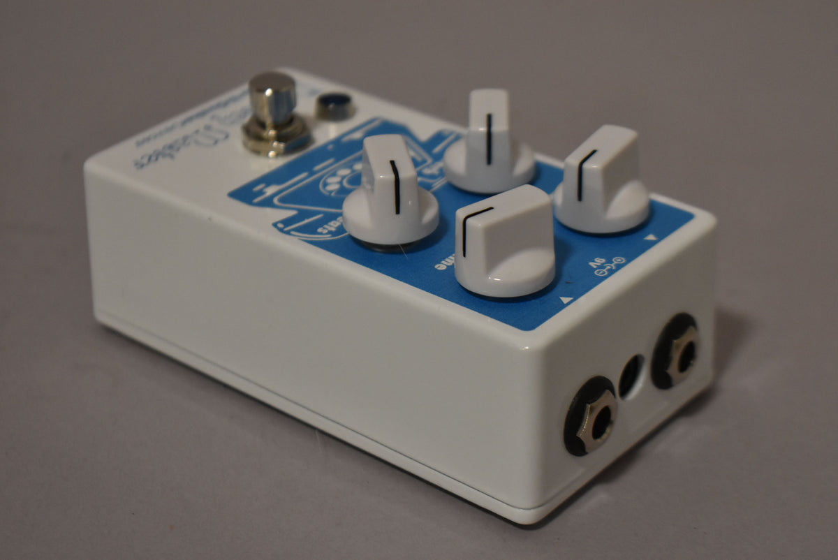 White effect pedal, angled from the front right, showing the top with four white knobs and a blue graphic, and the right side with two input jacks.