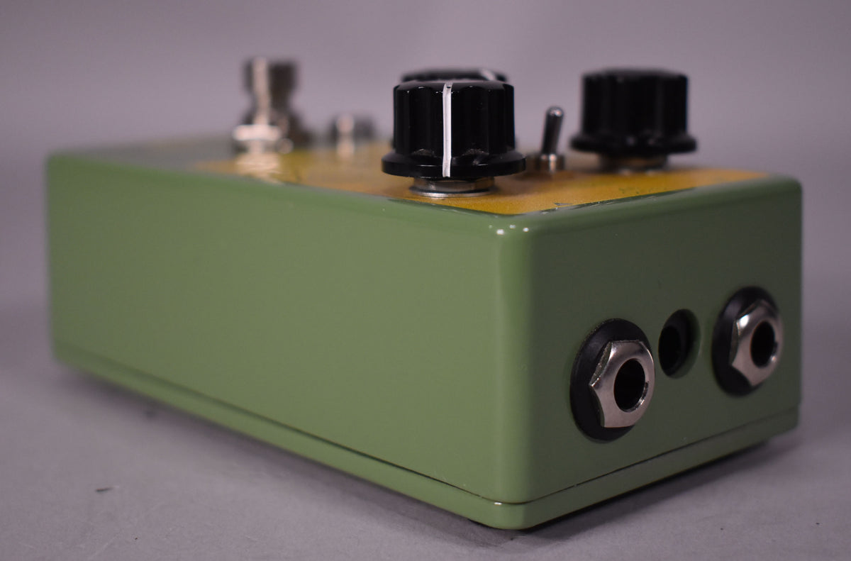 A green and yellow pedal, viewed from the right, showing two input jacks, a toggle switch, and two black control knobs.