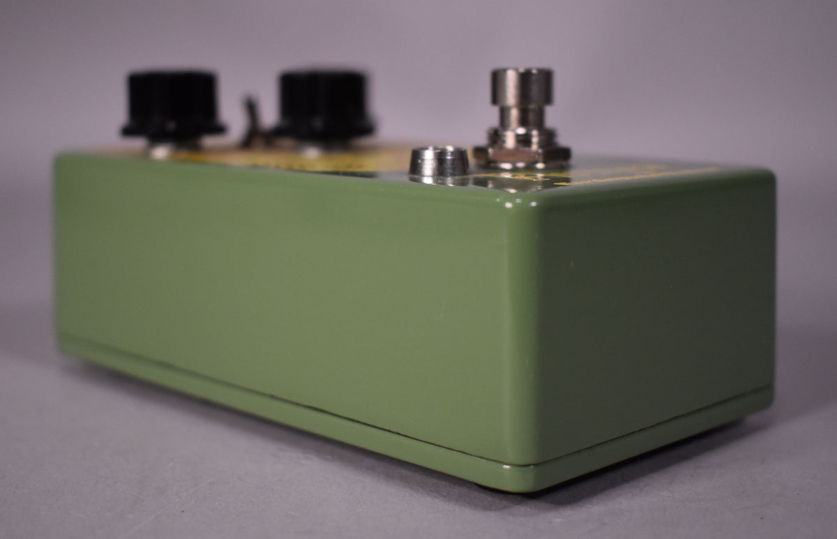 The olive green pedal is viewed from the right, showing two black knobs and a silver footswitch on its top surface.