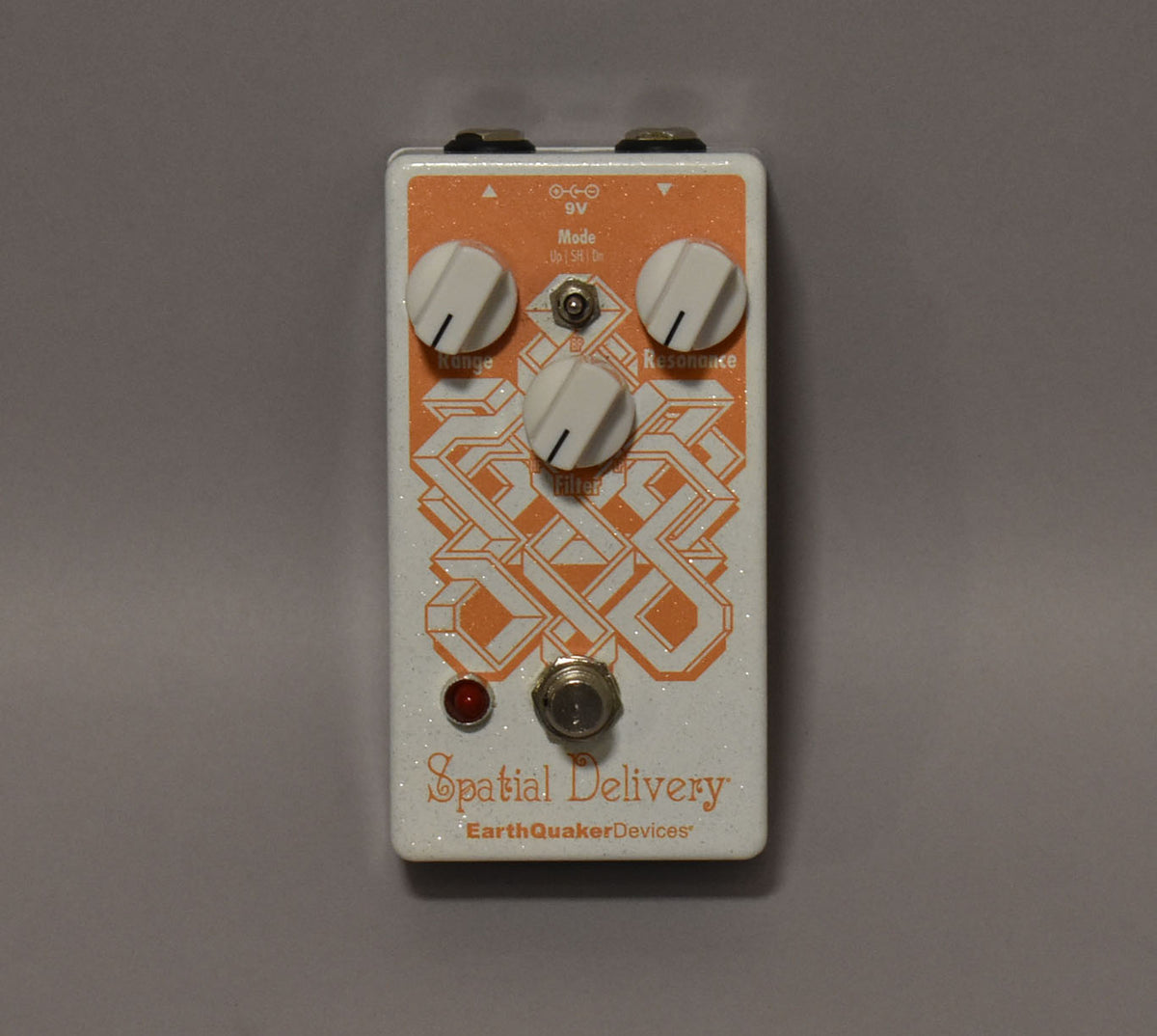 A white glitter-finish pedal with an orange geometric pattern, three white knobs, a small toggle switch, a red LED, and a footswitch.