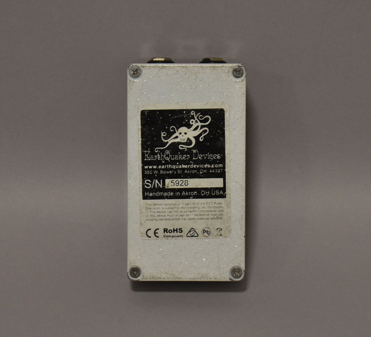 The white glitter-finish pedal is viewed from the back, showing a black label with an octopus logo and serial number.