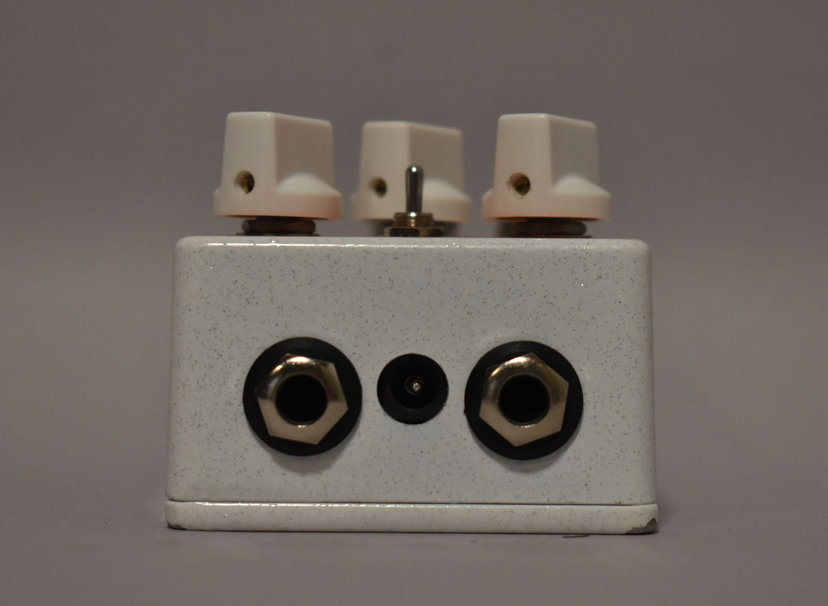 The white speckled pedal is viewed from the front, showing three cream knobs, a toggle switch, two input jacks, and a power input.