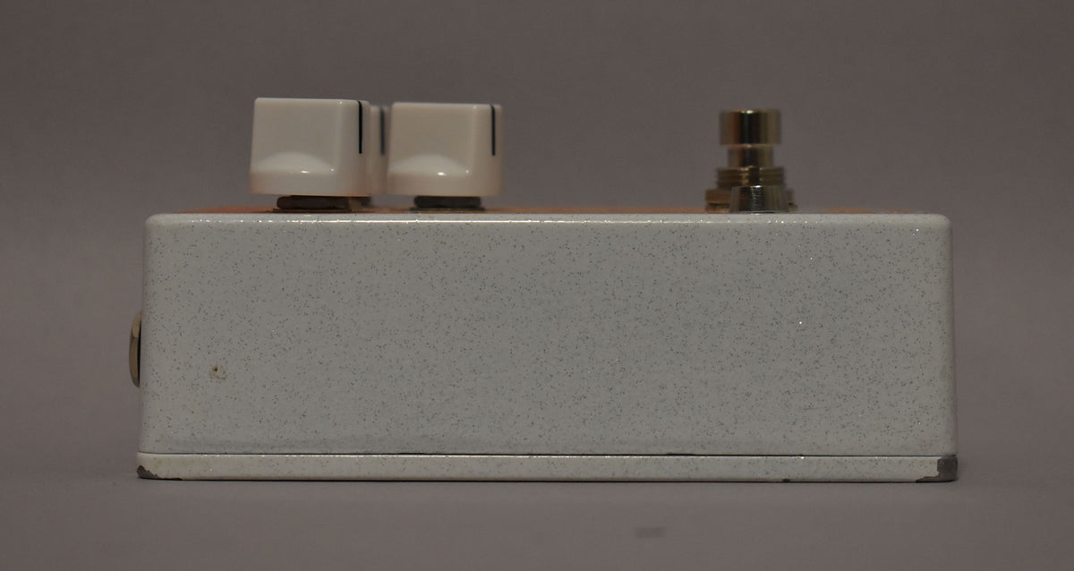 The white speckled pedal is seen from the right side, showing two white knobs and a silver footswitch on top, with some paint chips.