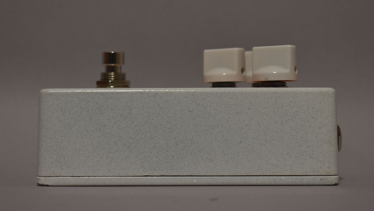 The white speckled pedal is viewed from the right side, showing a silver footswitch, two cream square knobs, and a silver input jack.