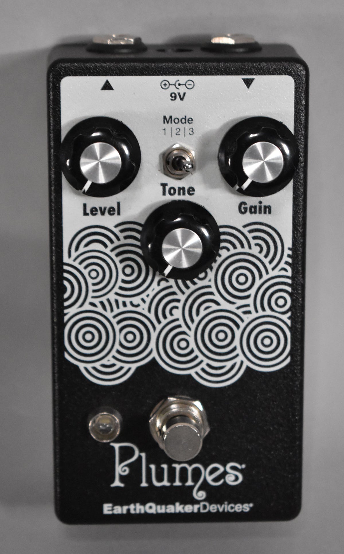 Black textured pedal with a white faceplate featuring a pattern of concentric circles, three black knobs, and a toggle switch.