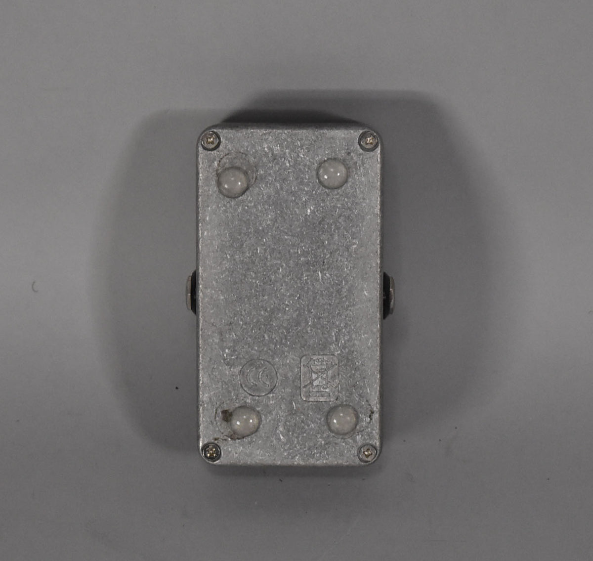 Bottom view of a rectangular, textured silver pedal with four round rubber feet and visible screw heads.