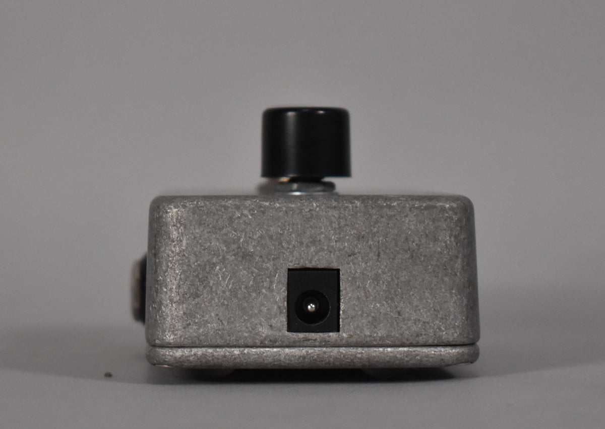 The pedal is viewed from the rear, showing the textured gray metal casing, a black power input jack, and a single black control knob on top.