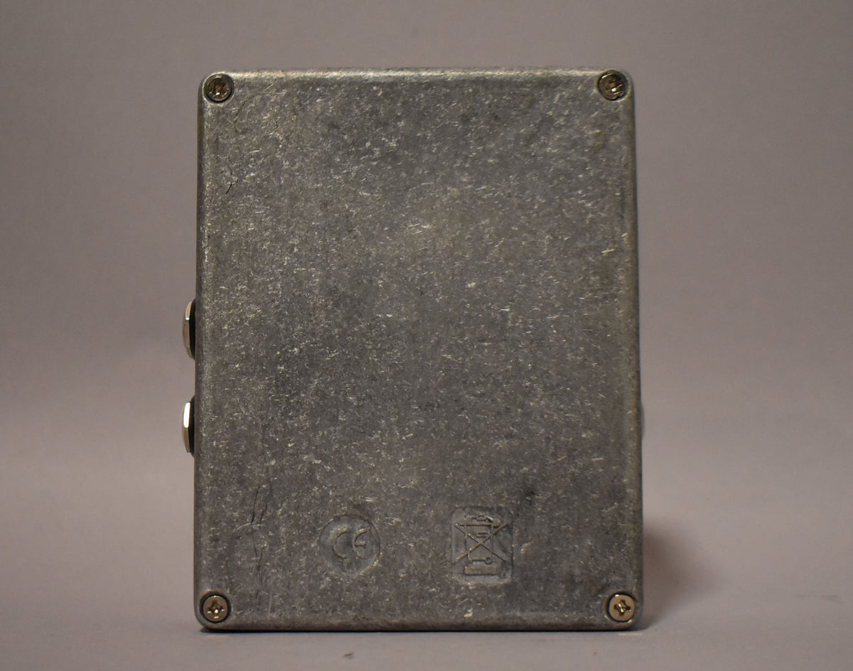 The pedal's textured gray metal casing is viewed from the bottom, showing four corner screws and two side jacks.