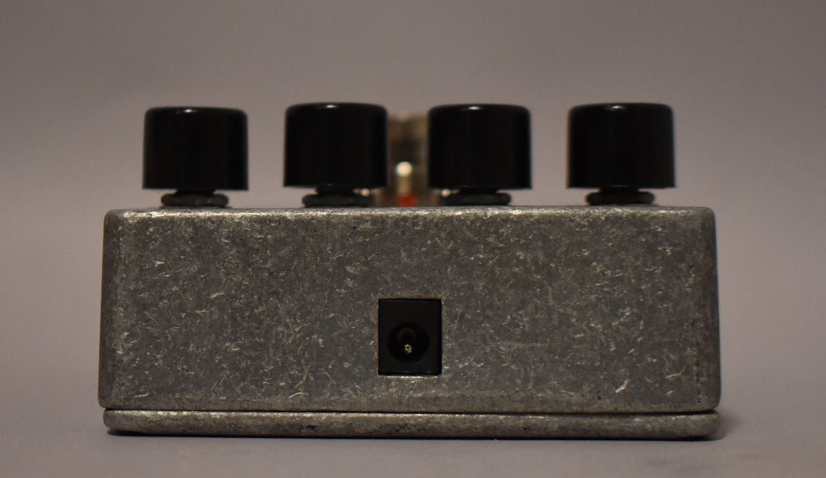 The effect pedal is viewed from the back, showing its textured gray metal casing, four black control knobs, and a central power input jack.