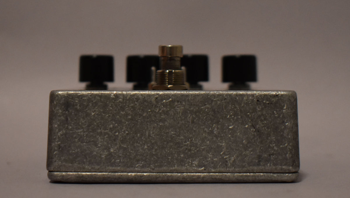 The pedal's textured gray metal casing is viewed from the side, with four black knobs and a brass footswitch visible on top.