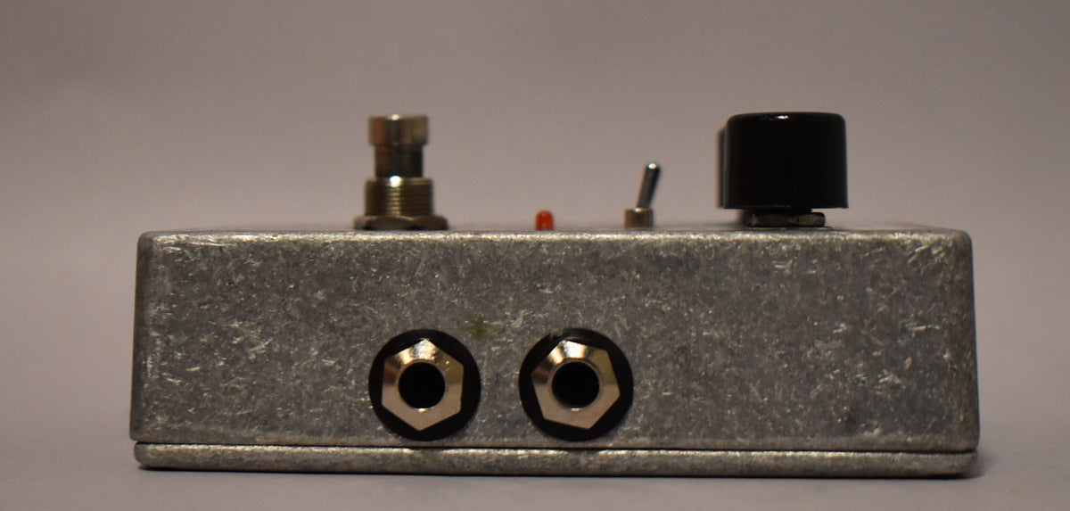The silver-gray pedal is viewed from the side, showing two input jacks, a footswitch, a red LED, a toggle switch, and a black knob.