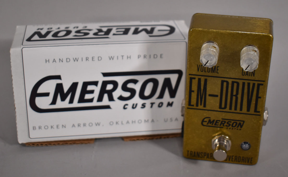 A gold effects pedal with clear Volume and Gain knobs, a silver footswitch, and a small blue LED, next to its white box.