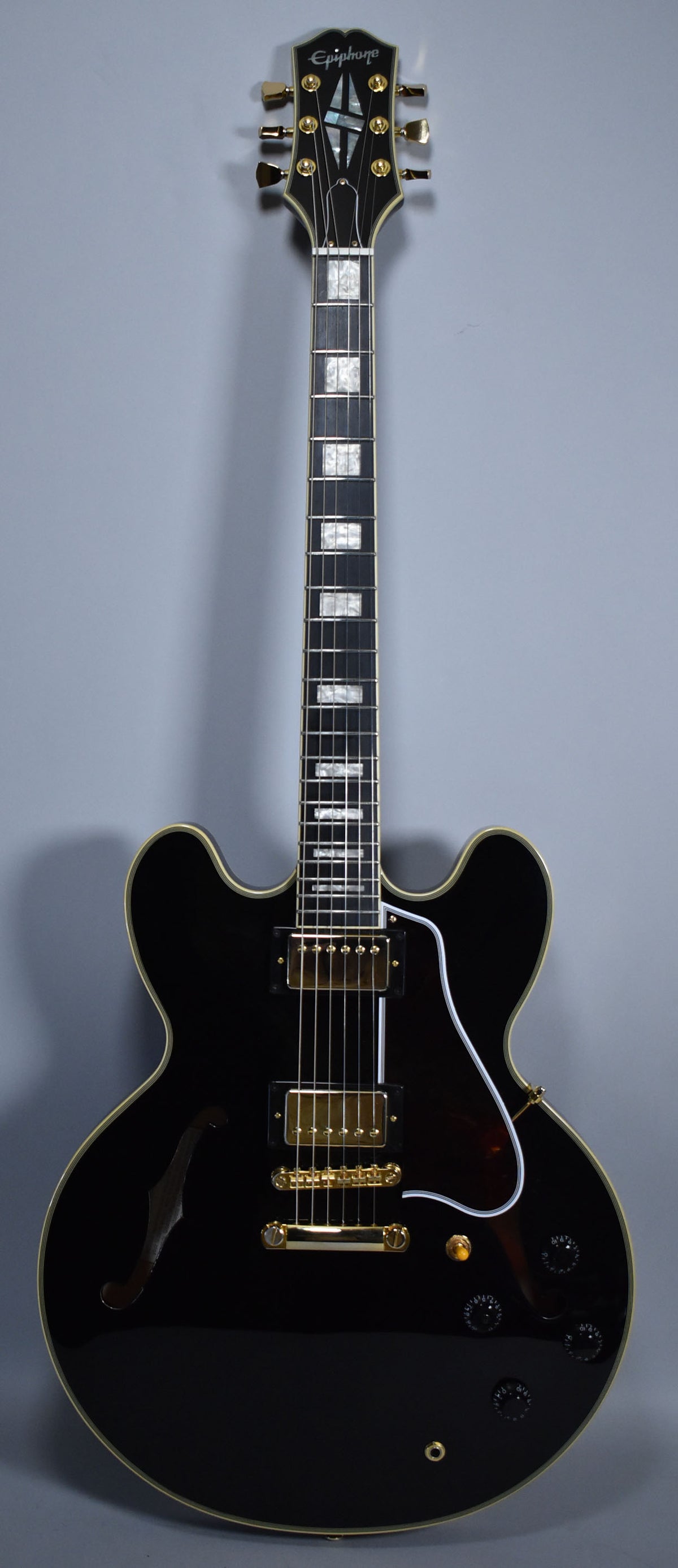 Full-length view of a glossy black semi-hollowbody guitar with gold hardware, white binding, and rectangular mother-of-pearl fretboard inlays.